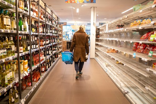 Persoon loopt tussen de schappen van een winkel met een winkelmandje in linkerhand.